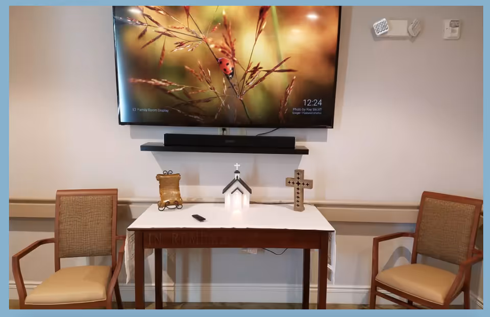A small memorial table with a white cloth, a miniature church model, a decorative cross, and a plaque. Two wooden chairs with cushioned seats are placed on either side of the table. Above the table is a mounted flat-screen TV displaying an image of a ladybug on a plant.
