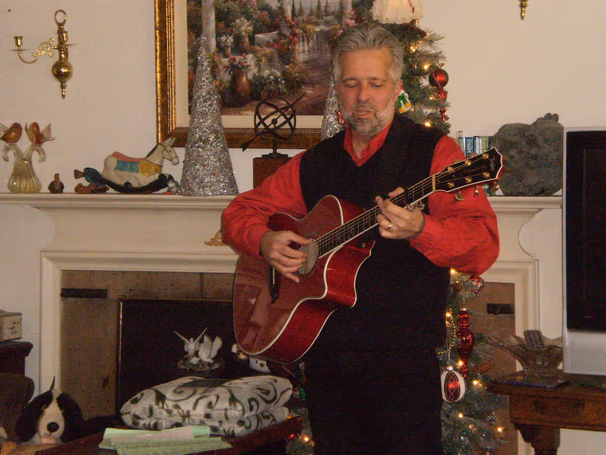 A man wearing a red shirt and black vest is playing an acoustic guitar in a cozy living room decorated for Christmas. Behind him is a fireplace mantel adorned with decorative items and a framed painting. A Christmas tree with ornaments and lights is visible to the right.
