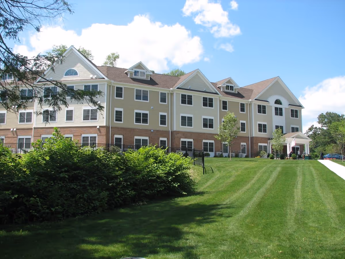 Exterior view of a three-story senior living facility building with beige and brick facade, multiple windows, a sloped roof with dormers, and a green lawn with bushes and trees under a partly cloudy blue sky.