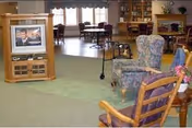 A spacious living room area in a senior living facility with carpeted floors, several upholstered armchairs, a wooden rocking chair, a wooden entertainment center with a television, and tables with chairs near large windows allowing natural light.