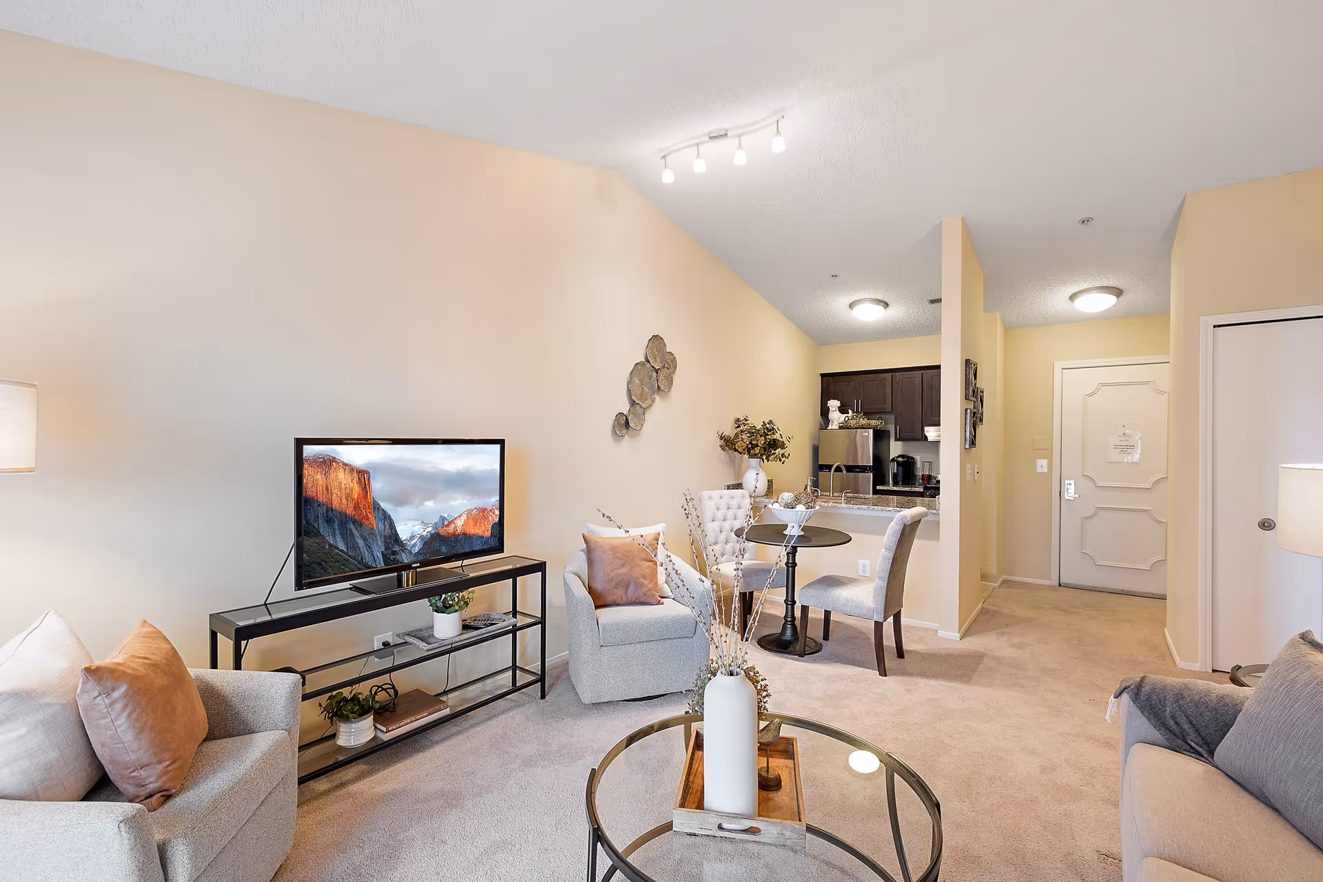 A cozy living room area with beige walls and carpet. The room features a TV on a black metal stand, two armchairs with cushions, a round glass coffee table with a decorative vase, and a small round dining table with two chairs. The kitchen area with dark cabinets and stainless steel appliances is visible in the background, along with a hallway and a closed door.