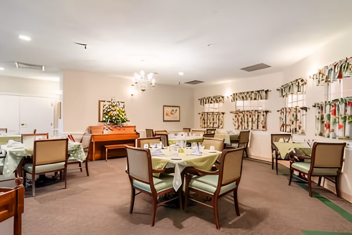 A dining room with multiple tables covered with light green tablecloths, each set with glasses and napkins. There are wooden chairs with light green cushions around the tables. A piano with a floral arrangement on top is against the far wall, and windows with decorative valances line one side of the room. The room is softly lit with ceiling lights and wall sconces.