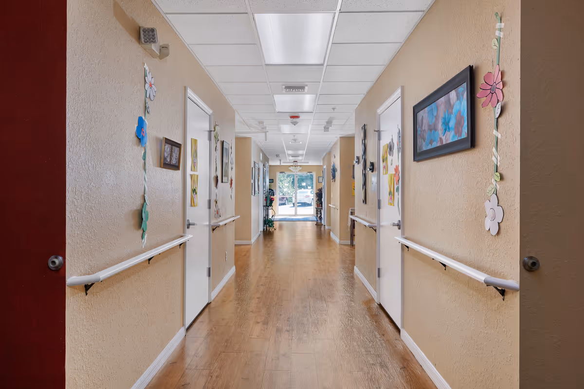 A well-lit hallway in an assisted living facility with wooden flooring, beige walls, white doors on both sides, handrails along the walls, and colorful flower decorations and framed artwork hanging on the walls. The hallway leads to a glass door with a view of the outside.