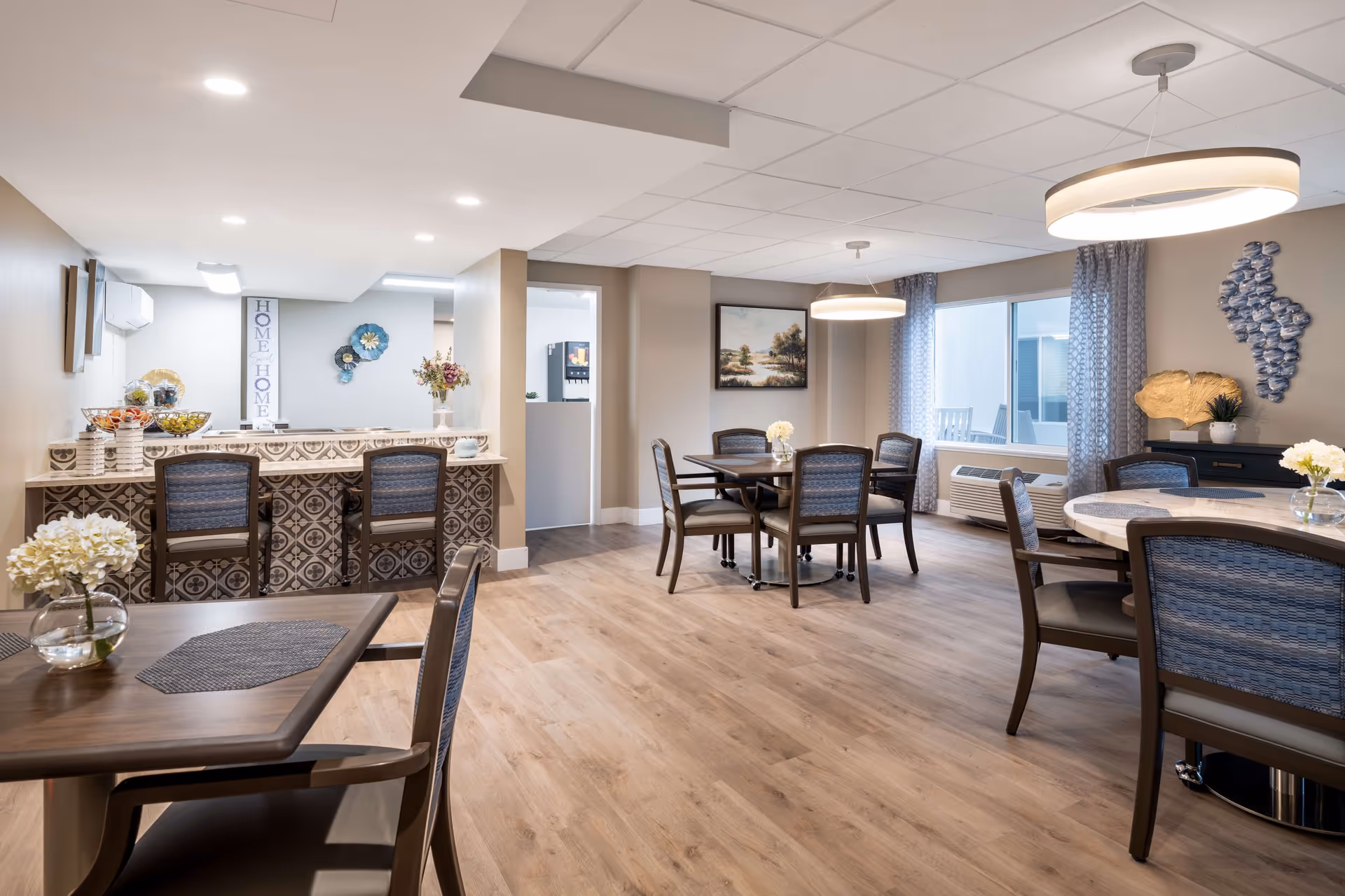 A bright and spacious dining area with multiple tables and chairs arranged for seating. The room features wood flooring, modern light fixtures, decorative wall art, and large windows with patterned curtains allowing natural light to enter. A counter with bar stools and decorative items is visible on the left side.