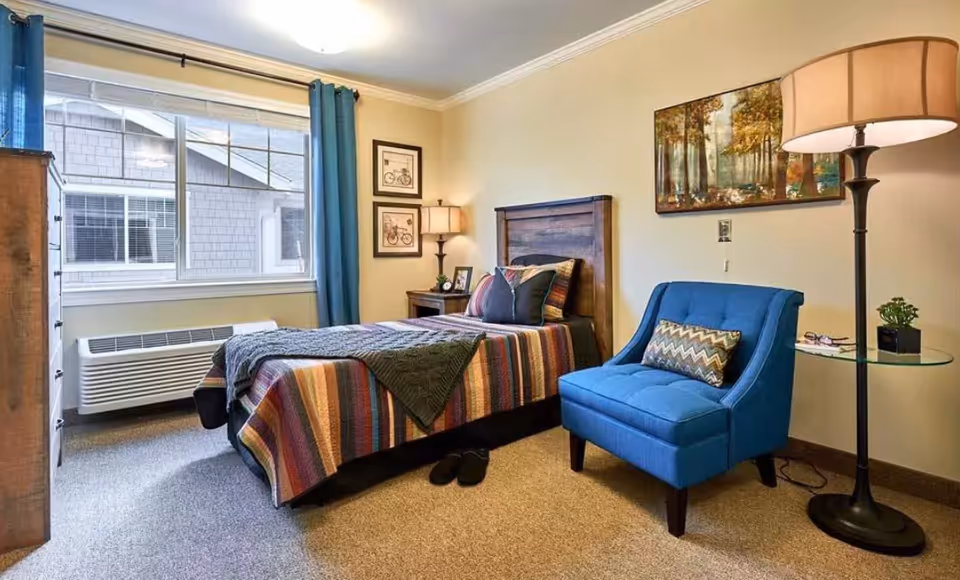 A cozy bedroom with a single bed covered in a colorful striped blanket and a dark green throw. Next to the bed is a wooden nightstand with a lamp, a small plant, and framed pictures on the wall. A blue upholstered chair with a patterned pillow sits beside a round glass side table holding a small plant and a pair of glasses. A tall floor lamp stands next to the chair. The room has beige walls, a large window with blue curtains, and a carpeted floor.