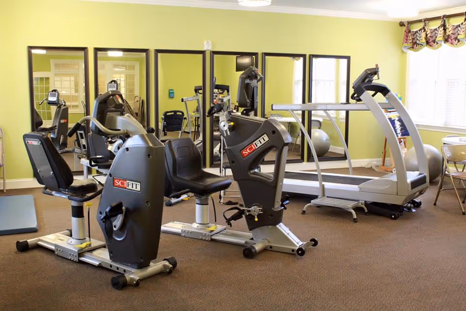 Small fitness room with recumbent exercise bikes, a treadmill, mirrors on the wall, and a window with curtains.