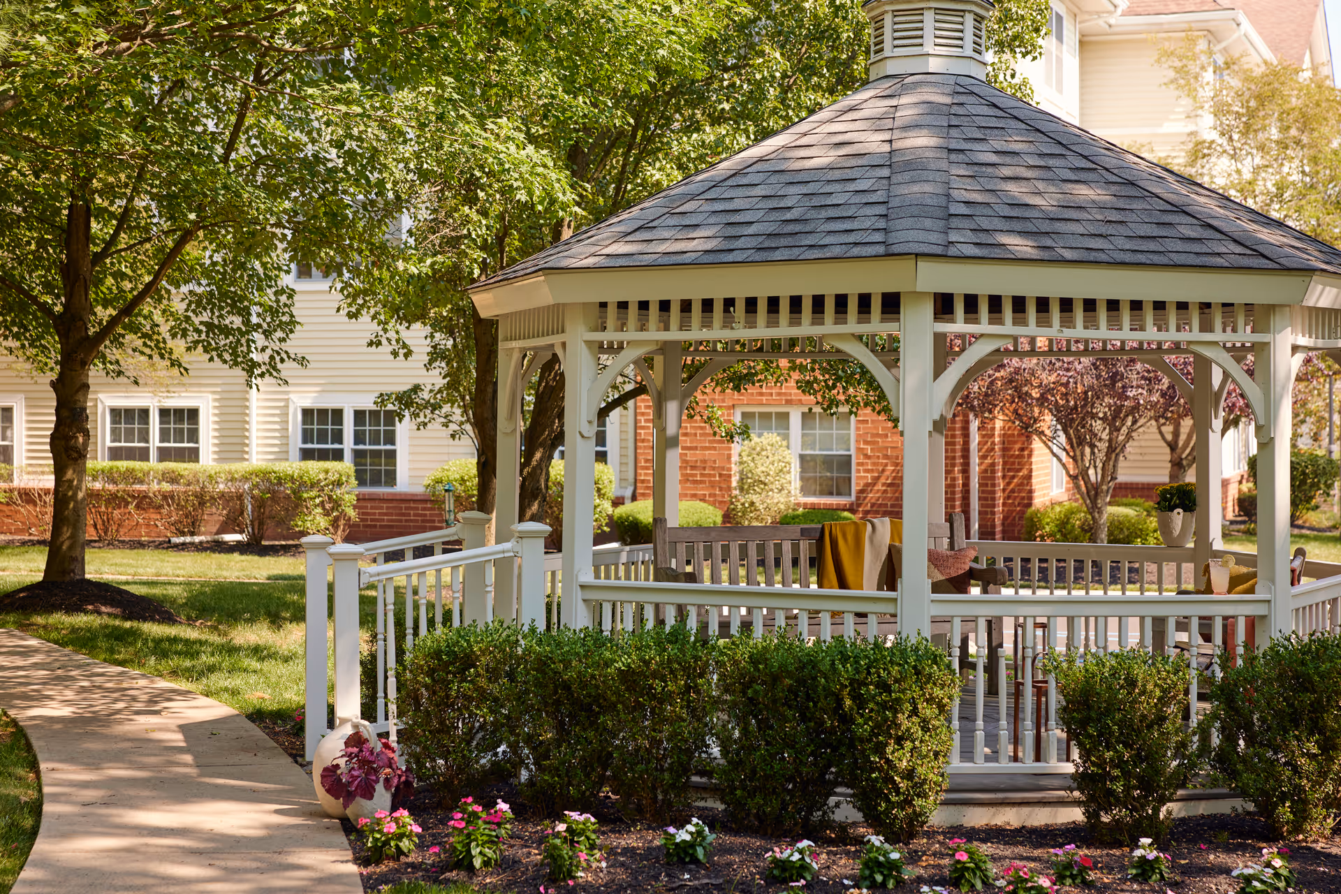 A white wooden gazebo with a shingled roof situated in a garden area with green bushes, colorful flowers, and trees. There are benches inside the gazebo with a blanket draped over one of them. In the background, there is a multi-story building with beige siding and red brick accents.