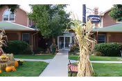 Exterior view of Cinnamon Park facility entrance with a concrete walkway flanked by green lawns, bushes, and autumn decorations including pumpkins and cornstalks. The building has a brown exterior with a green roof and a sign that reads 'Cinnamon Park'.