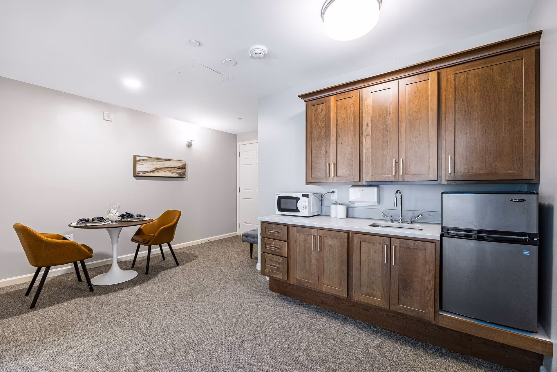 Small kitchenette with wooden cabinets, a sink, microwave and mini-fridge beside a round dining table with two mustard-colored chairs.