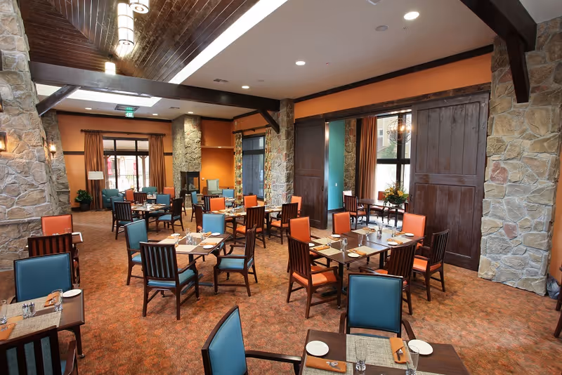 A spacious dining room with multiple tables set for meals, featuring chairs with blue and orange cushions. The room has stone pillars, warm orange walls, large windows with curtains, and a cozy fireplace in the background. The ceiling has wooden beams and modern light fixtures.