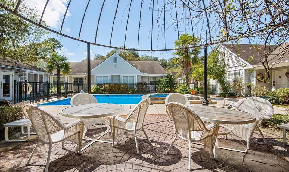 Patio with wicker tables and chairs under a metal pergola beside a swimming pool, surrounded by single-story residential buildings and landscaping.