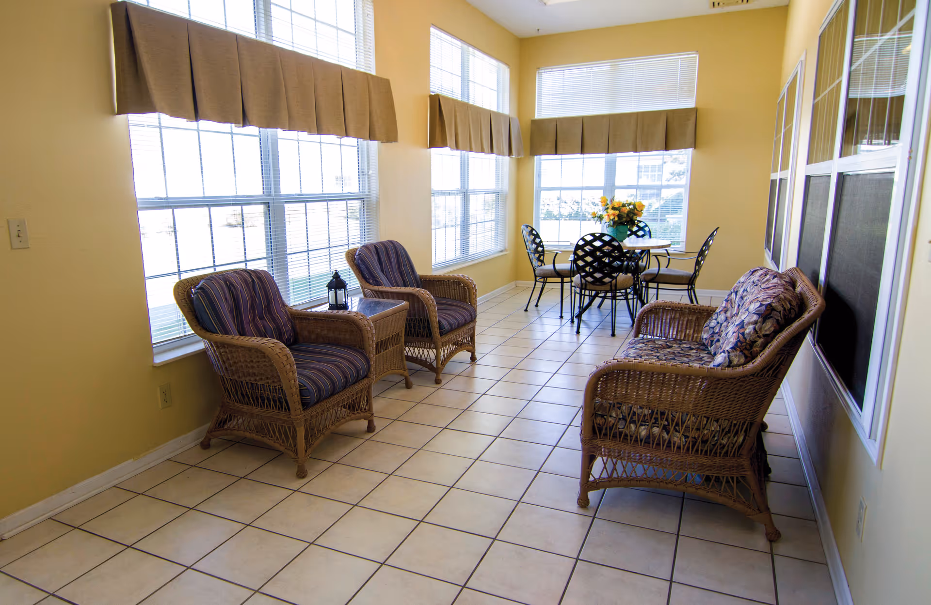 Sunlit seating area with wicker chairs and a small dining table near large windows in a tiled room.