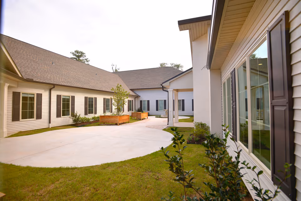 Outdoor courtyard area of a senior living facility with beige siding buildings, multiple windows with dark shutters, a concrete walkway, green grass, and planter boxes with small trees and shrubs.