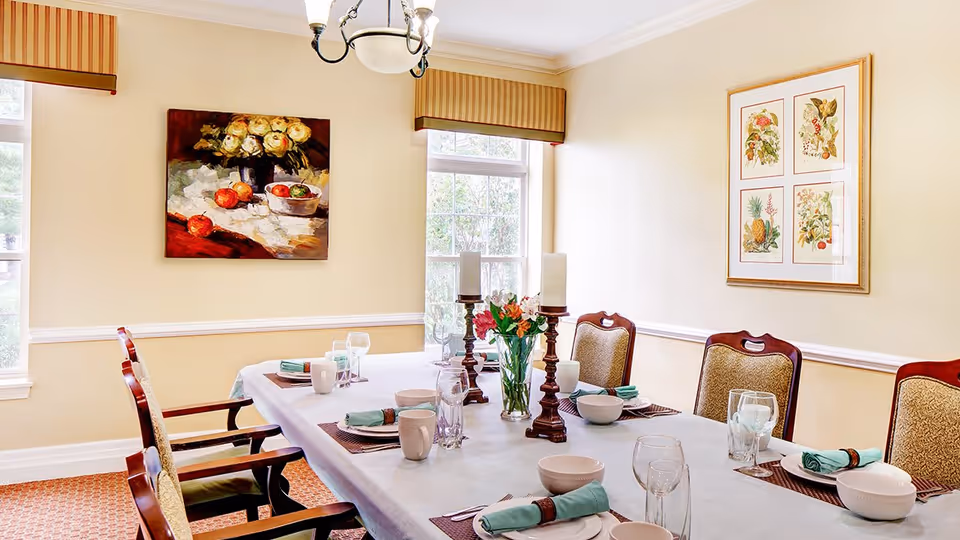 A well-lit dining room with a rectangular table set for six people. The table is covered with a white tablecloth and has place settings including bowls, mugs, glasses, and napkins with napkin rings. There are two tall candle holders with white candles and a vase with flowers in the center. The room has beige walls with two windows, framed artwork on the walls, and a chandelier hanging from the ceiling.