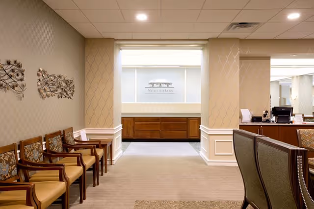 A waiting area in a senior living facility with a row of wooden chairs with patterned cushions along the left wall, decorative metal wall art above the chairs, and a reception desk with chairs on the right. The area is well-lit with recessed ceiling lights and has beige walls with white wainscoting. At the far end, there is a frosted glass panel with the North Oaks logo and name.