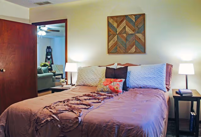 A bedroom with a neatly made bed featuring a mauve bedspread, decorative pillows, and a wooden geometric wall art above the headboard. Two bedside tables with lamps flank the bed. An open door reveals a glimpse of a living room area with a sofa and a ceiling fan.