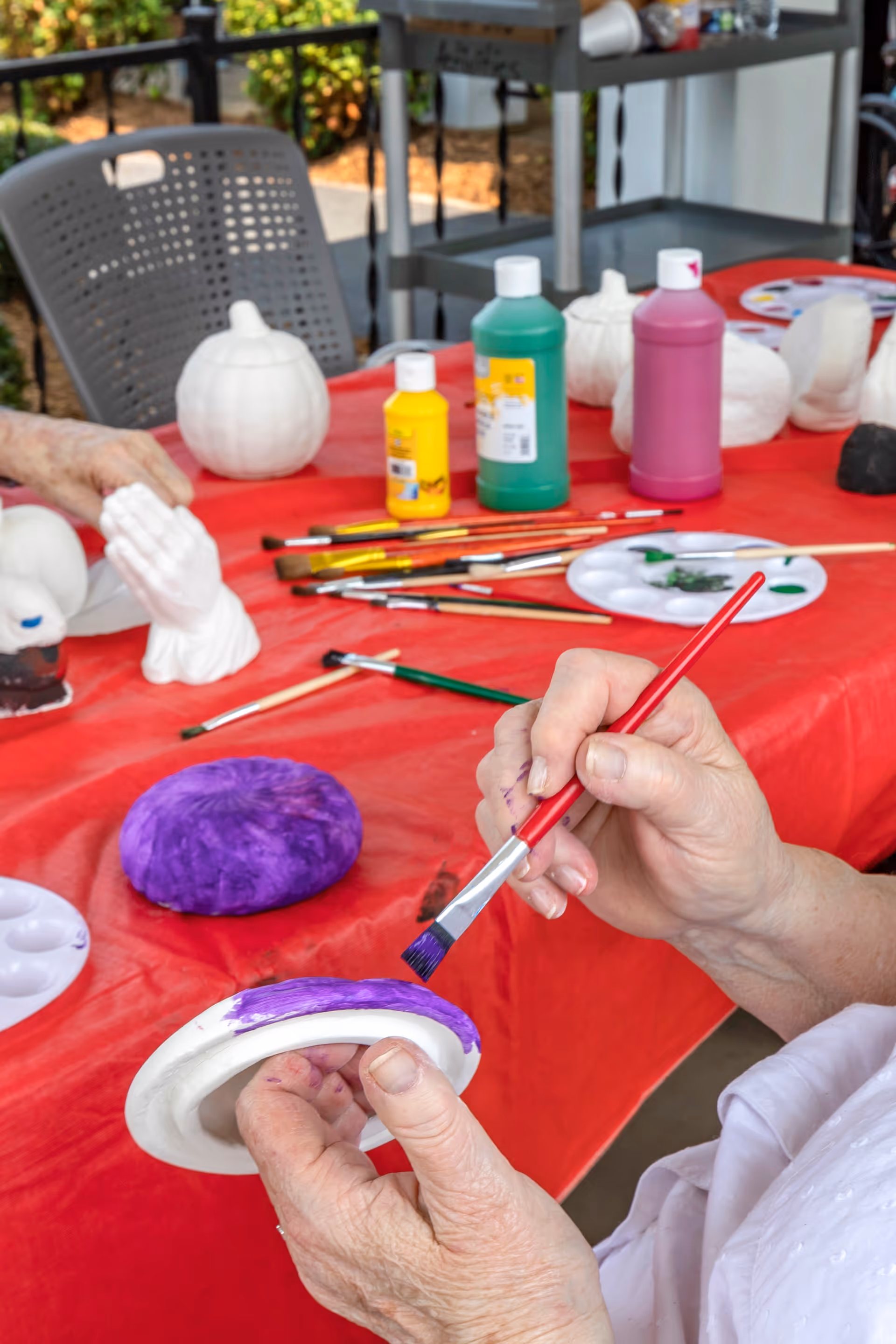 An elderly person painting a small white ceramic pumpkin with purple paint using a paintbrush at a table covered with a red tablecloth. Various paint bottles, brushes, and other ceramic pumpkins are visible on the table. The setting appears to be an outdoor or semi-outdoor craft activity area.