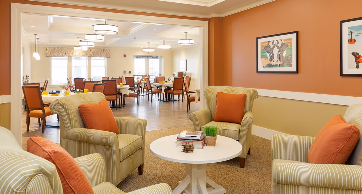 Cozy lounge area with striped armchairs and orange pillows around a white coffee table, opening into a dining room with tables and chairs.