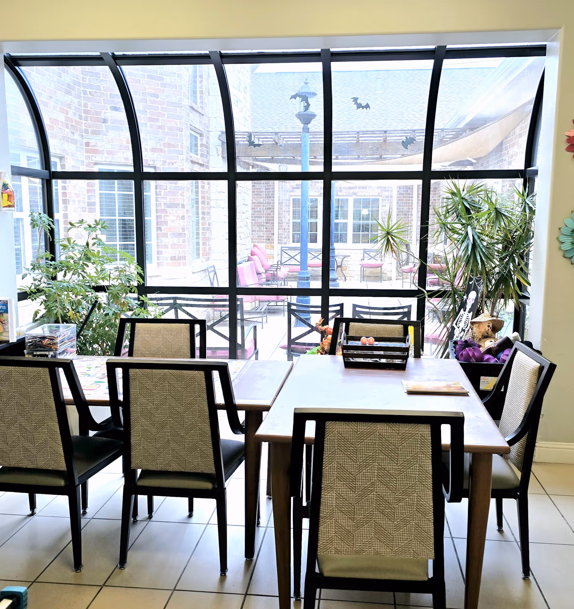 Indoor seating area with several chairs and tables arranged near a large curved glass window that looks out onto a patio with more seating and plants. The room has potted plants and some Halloween decorations including a skeleton figure and bat stickers on the window.