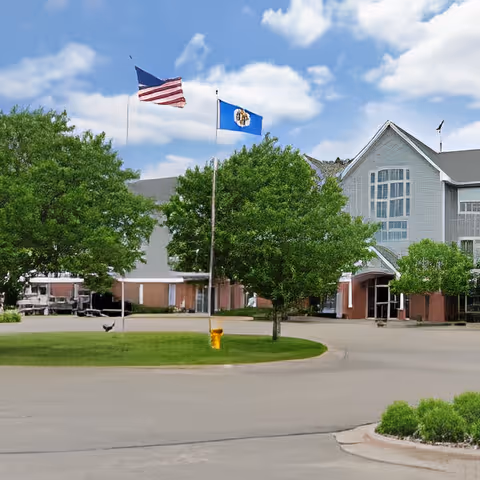 Exterior view of Carefree Cottages of Maplewood facility with two flagpoles displaying the American flag and a state flag, surrounded by green trees and a circular driveway under a partly cloudy sky.