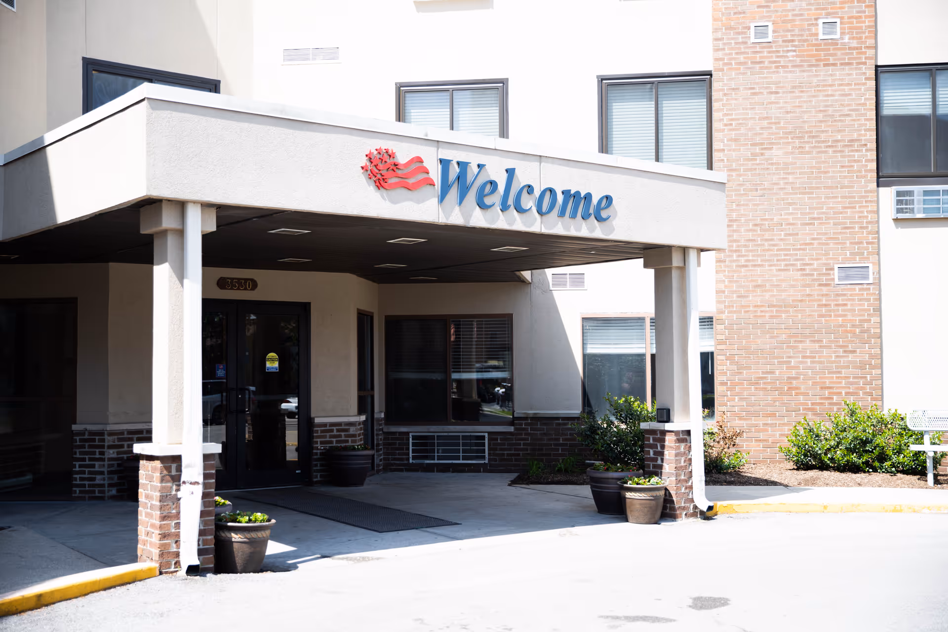 Entrance of a building with a covered drop-off area featuring a sign that says 'Welcome' with a red and blue flag design above it. The building has brick and beige walls, windows with blinds, and several potted plants near the entrance.