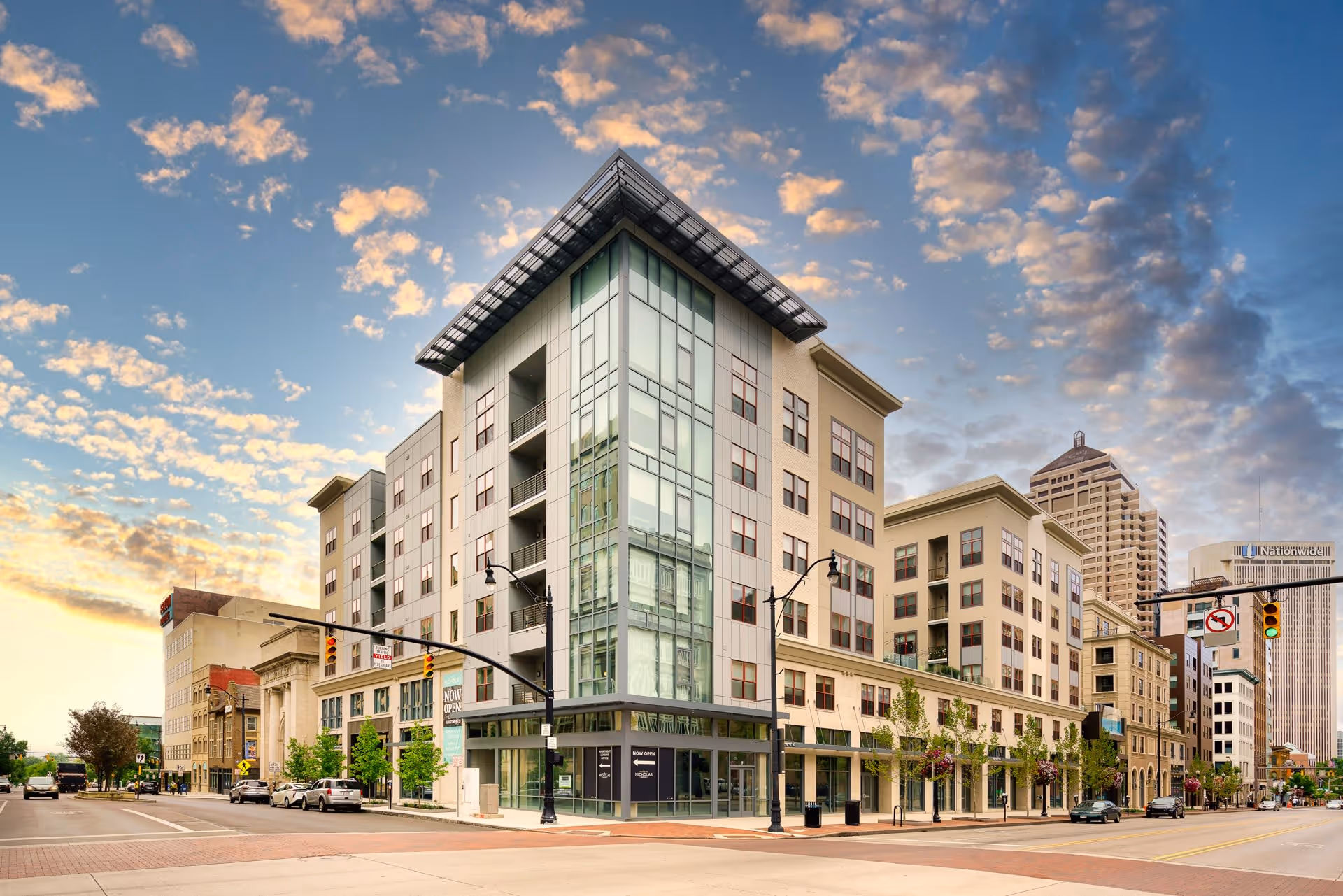 Modern multi-story building with large glass windows on a city street corner under a partly cloudy sky during sunset, with several cars parked along the street and other buildings in the background.