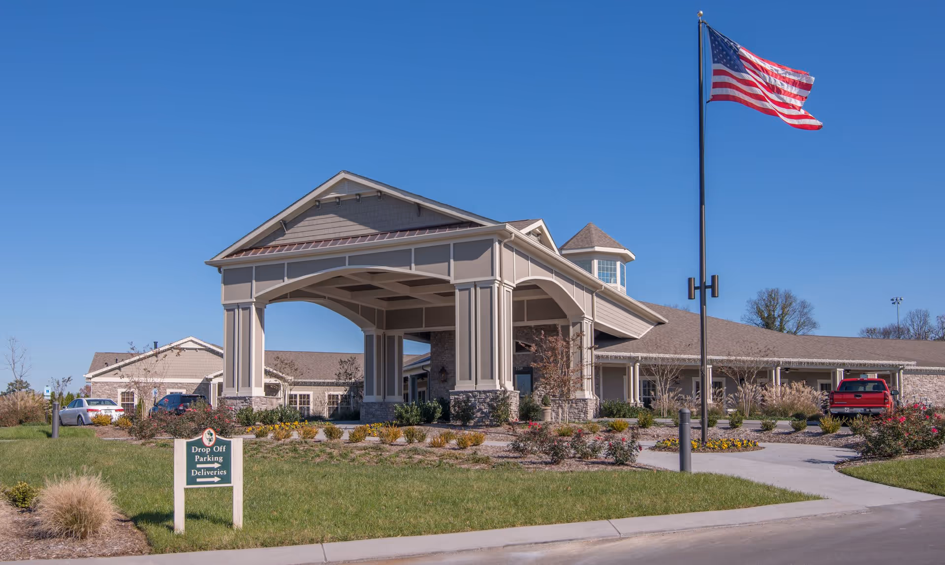 Front entrance of a senior living facility with a covered porte-cochere, landscaped grounds, parked cars, and an American flag on a tall pole.