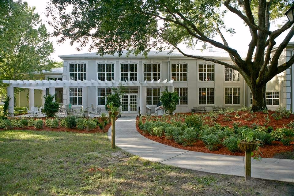 Exterior view of The Gables of Palm Harbor building with large windows, a white pergola, outdoor seating, landscaped garden beds with shrubs and mulch, a concrete walkway, and a large tree providing shade.
