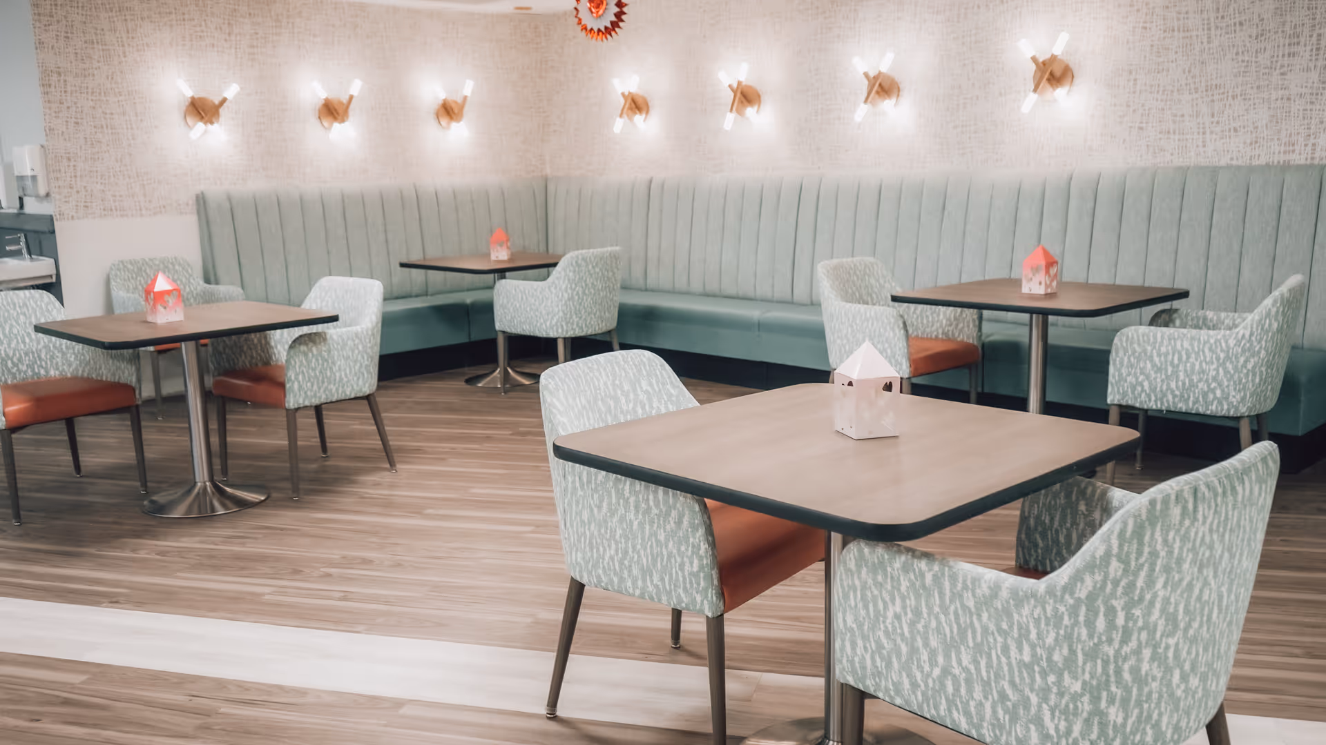 Modern dining area with square tables, upholstered chairs, and banquette seating under wall sconces.