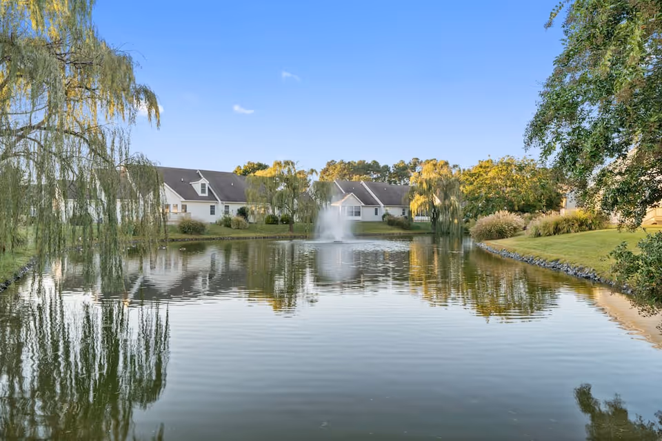 A tranquil pond with a central fountain surrounded by lawns, weeping willow trees, and single-story residential buildings reflected in the water.