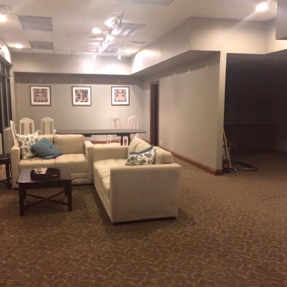 A cozy living room area with two beige sofas arranged around a dark wooden coffee table. The sofas have decorative pillows in blue and floral patterns. Behind the sofas is a dining table with white chairs and three framed artworks on the wall. The room has beige carpet and neutral-colored walls with ceiling lights.