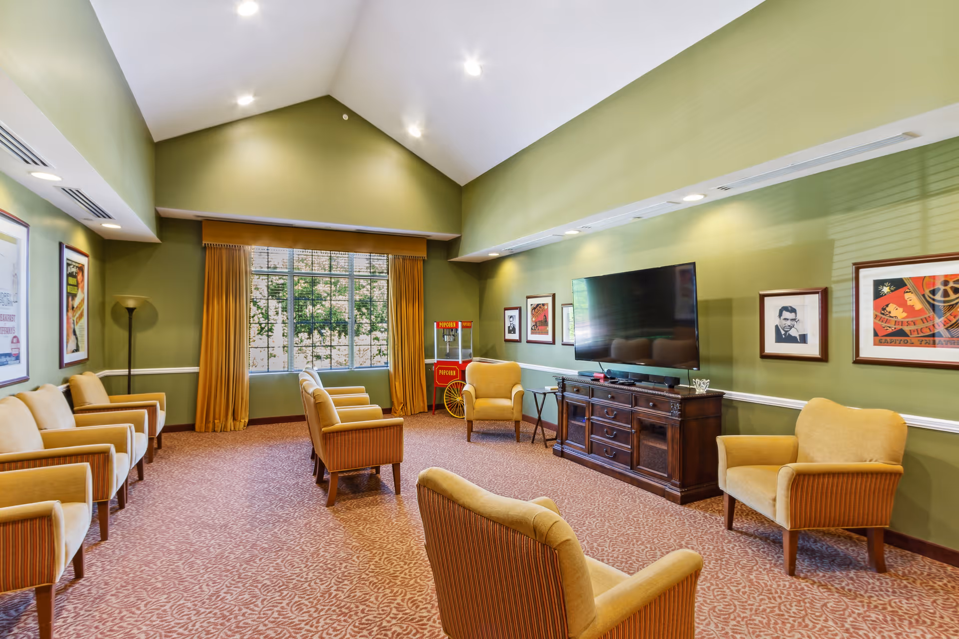 A spacious living room with green walls and a high vaulted ceiling. The room is furnished with multiple yellow upholstered armchairs arranged around a large flat-screen TV mounted on a wooden cabinet. There are framed pictures on the walls and a popcorn machine near a large window with yellow curtains, allowing natural light to enter.
