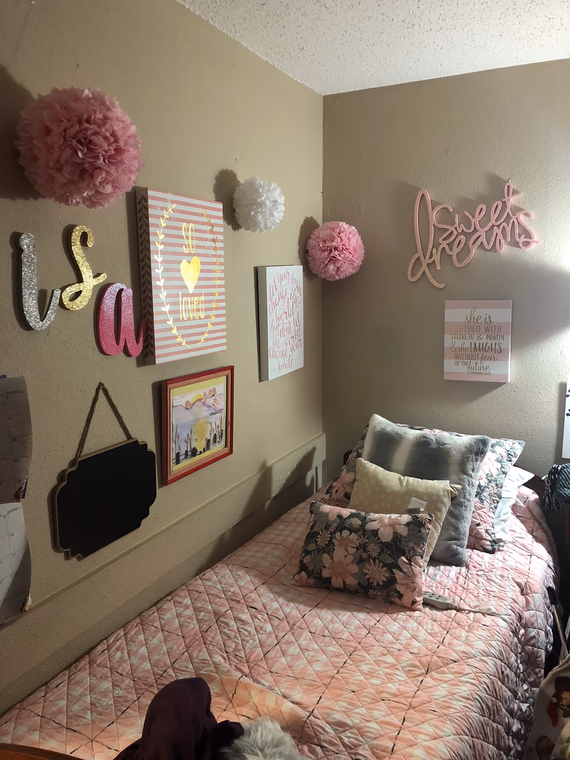 A cozy bedroom corner with a single bed covered in a pink quilt and multiple decorative pillows. The beige walls are adorned with various wall decorations including pink and white pom-poms, framed inspirational quotes, and a pink 'Sweet dreams' sign.