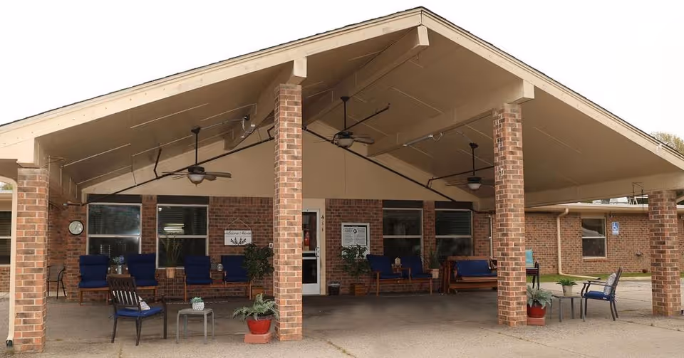 Covered entrance of a brick nursing home with a large gabled portico supported by brick columns, outdoor seating, ceiling fans, and potted plants.