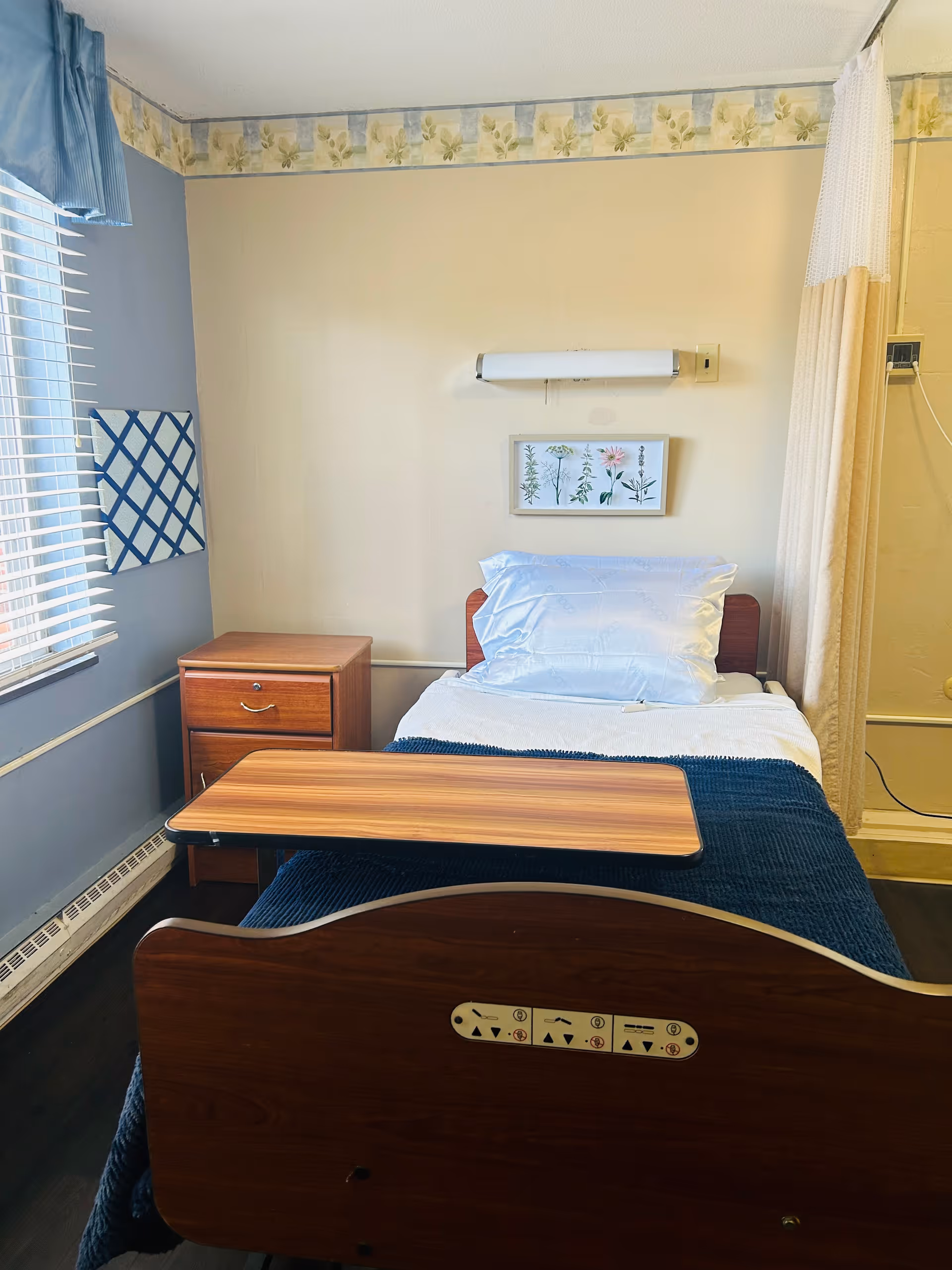 A single hospital-style bed with a wooden headboard and footboard in a small room. The bed is made with white sheets, a blue blanket, and a white pillow. A wooden overbed table is positioned over the bed. To the left of the bed is a wooden nightstand with two drawers. The room has a window with white blinds and a blue valance. The walls are painted beige and blue with a floral wallpaper border near the ceiling. A framed botanical print hangs above the bed, and a privacy curtain is partially visible on the right side.