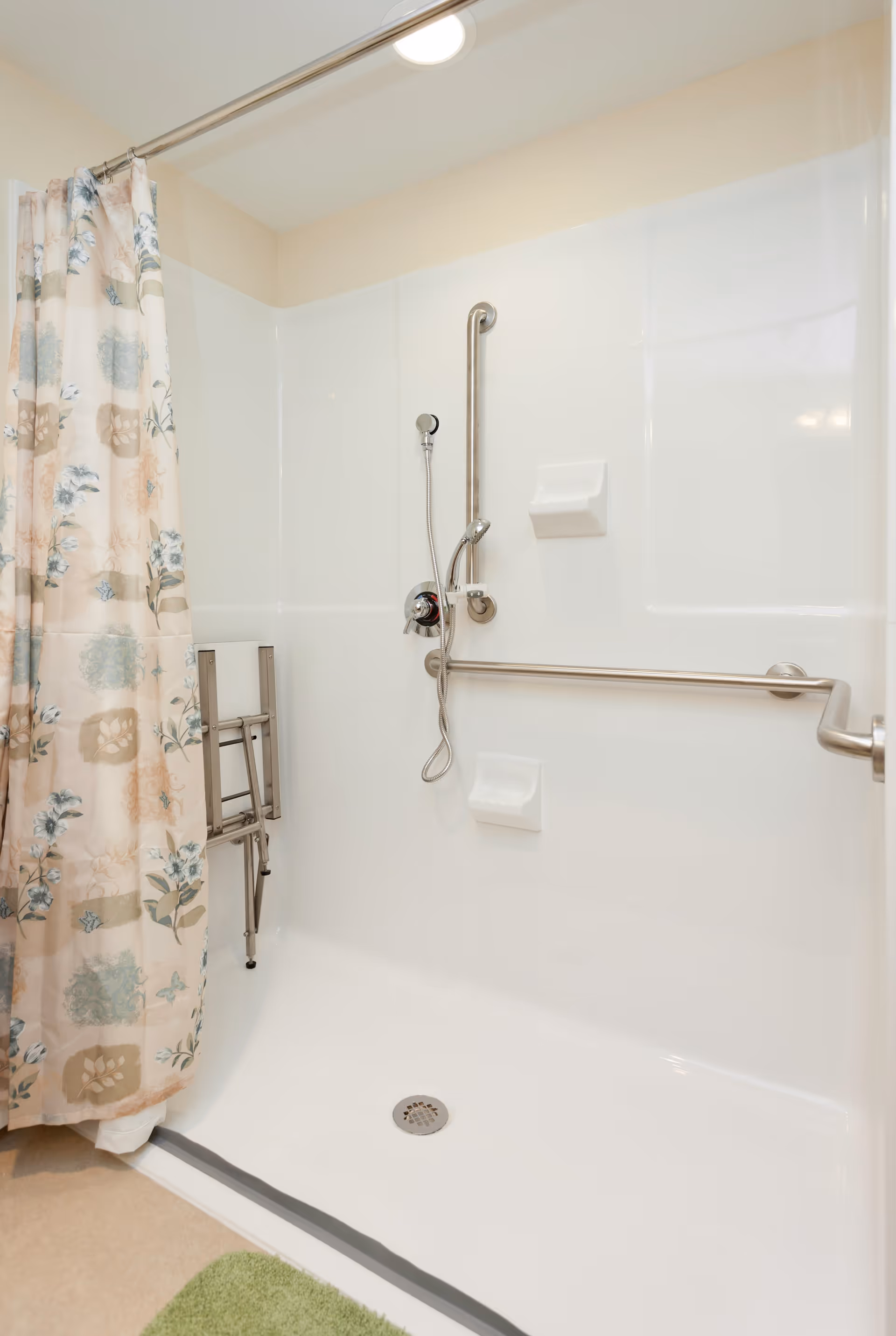 A clean, accessible shower area with a floral patterned shower curtain, a foldable shower chair, grab bars on the wall, a handheld showerhead, and soap holders.