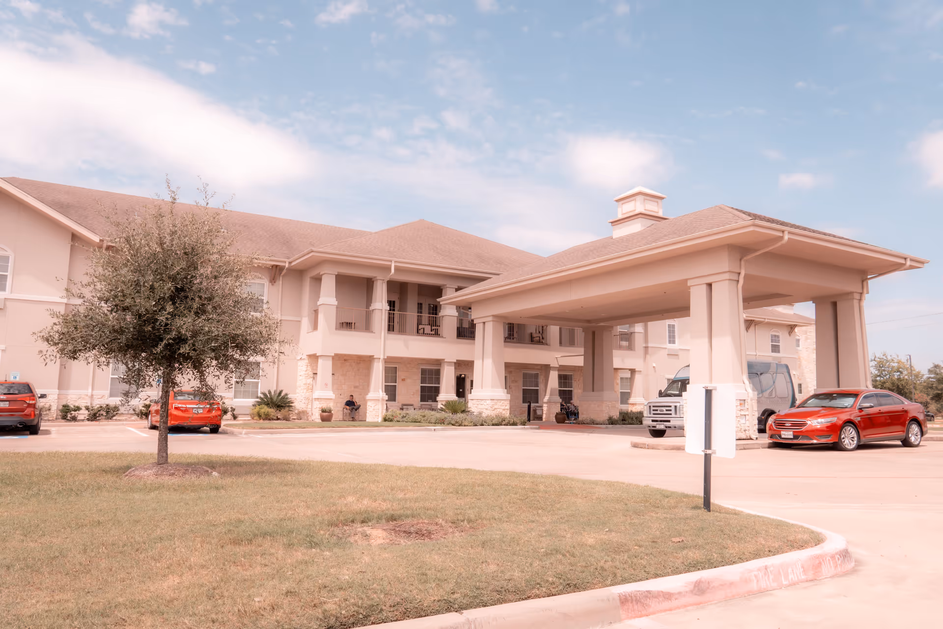 Exterior view of The Legacy at Falcon Point senior living facility showing a two-story building with a covered entrance, several parked cars, a small tree, and a clear sky with some clouds.