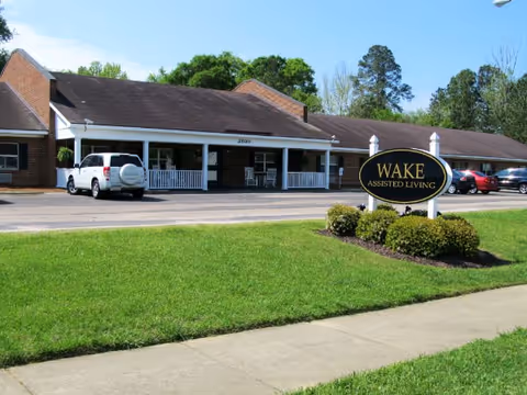 Front exterior of the Wake Assisted Living memory care building with a lawn, driveway, parked cars, and a sign reading "WAKE ASSISTED LIVING".