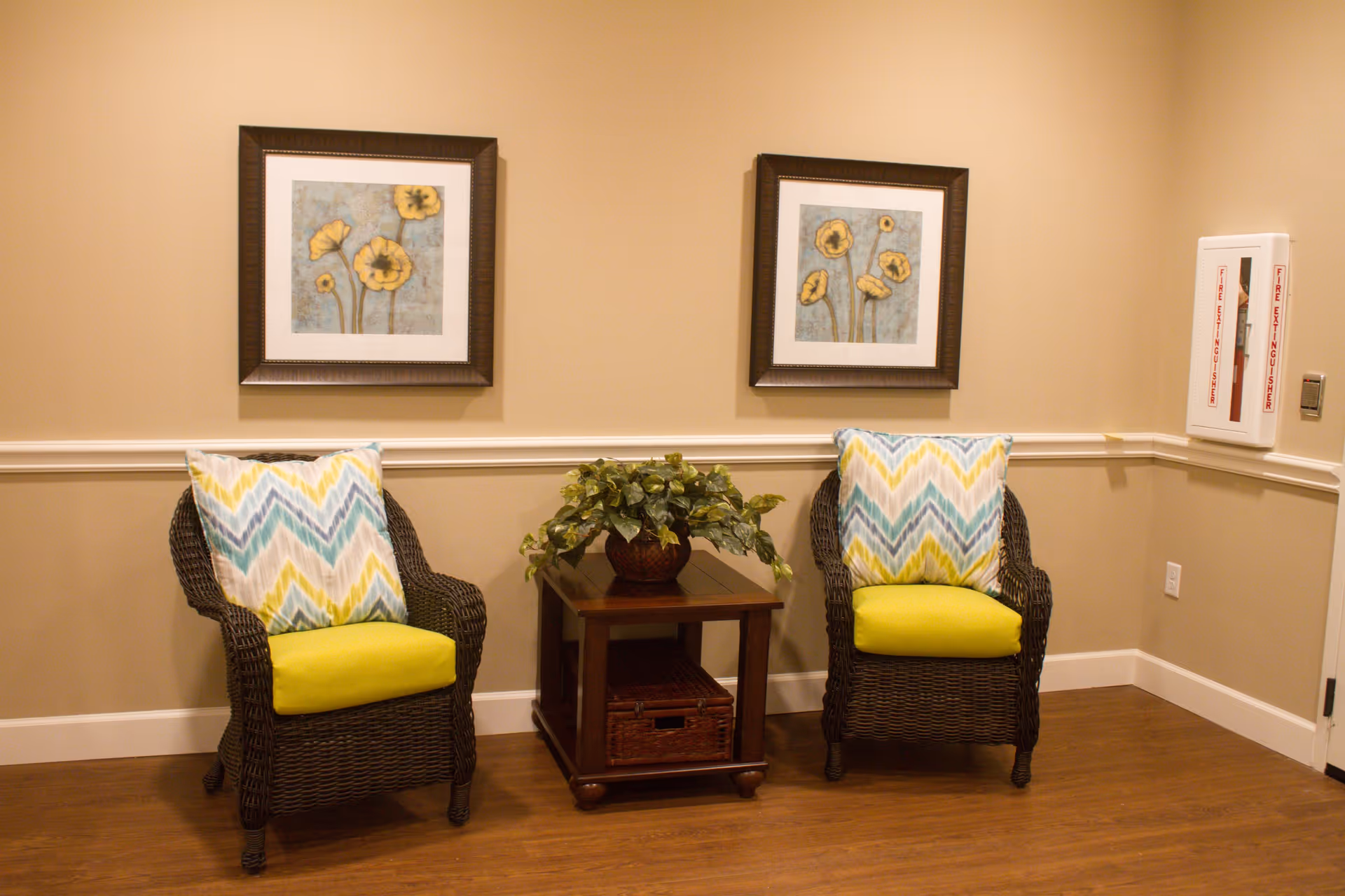 A cozy seating area with two wicker chairs featuring yellow cushions and chevron-patterned pillows in blue, yellow, and white. Between the chairs is a wooden side table with a potted green plant. Two framed floral paintings hang on the beige wall above the chairs. A fire extinguisher cabinet is mounted on the right wall.