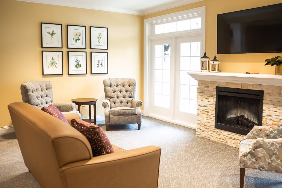 A cozy living room with beige walls, a tan sofa with purple patterned pillows, two tufted gray armchairs, a small round wooden side table, a stone fireplace with two decorative lanterns on the mantel, a flat-screen TV mounted above the fireplace, and a large windowed door letting in natural light. Six framed botanical prints hang on the wall.