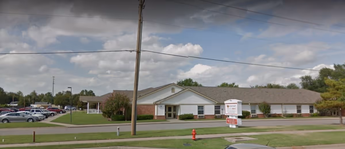 Exterior view of a single-story assisted living and memory care facility building with a parking lot on the left side, a green lawn, and a sign in front that reads 'Sommerset Neighborhood Assisted Living and Memory Care'. The sky is partly cloudy.