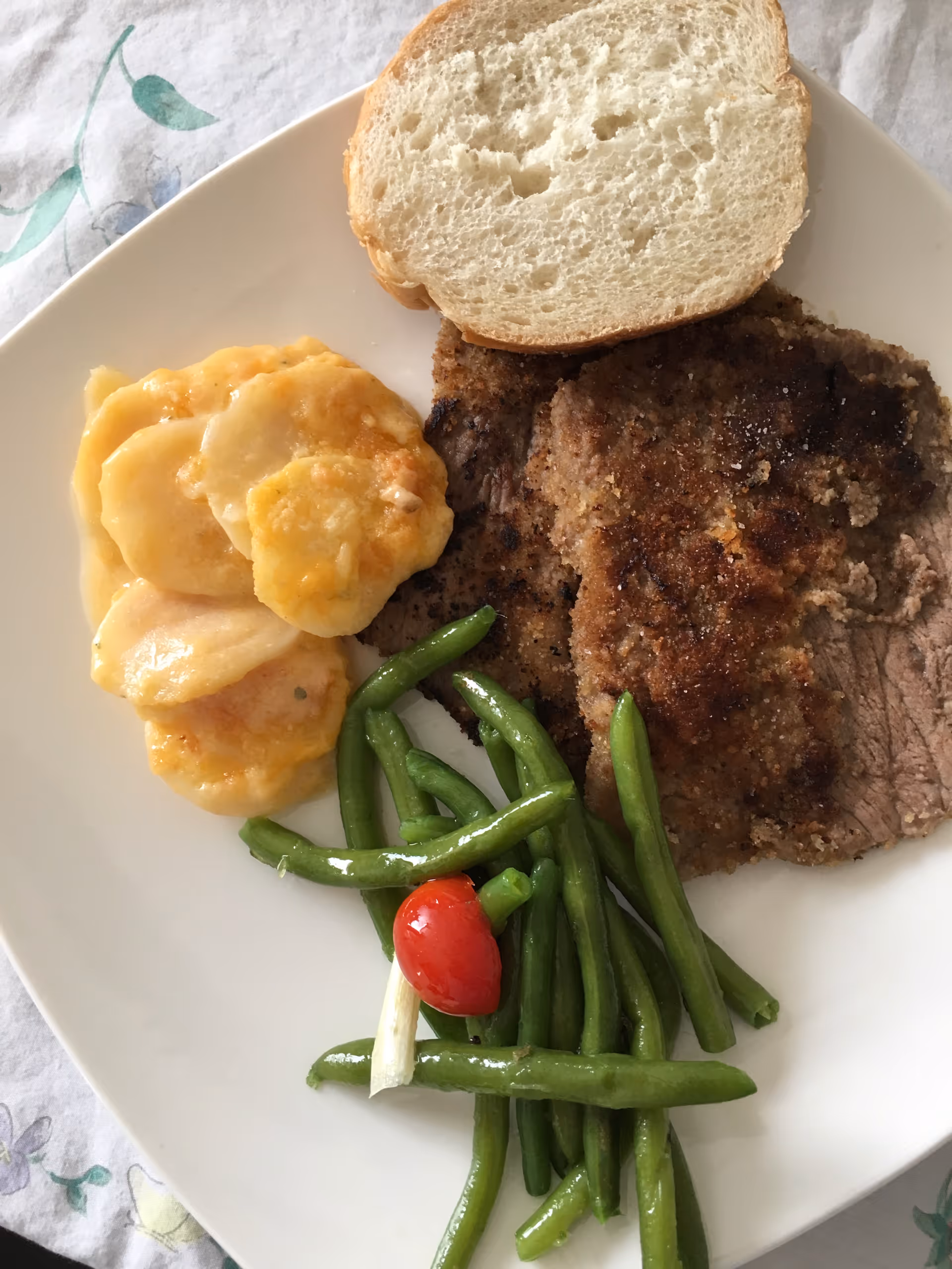 A plate of food with two pieces of breaded meat, a serving of scalloped potatoes with melted cheese, steamed green beans, and a small red cherry tomato on a white plate.