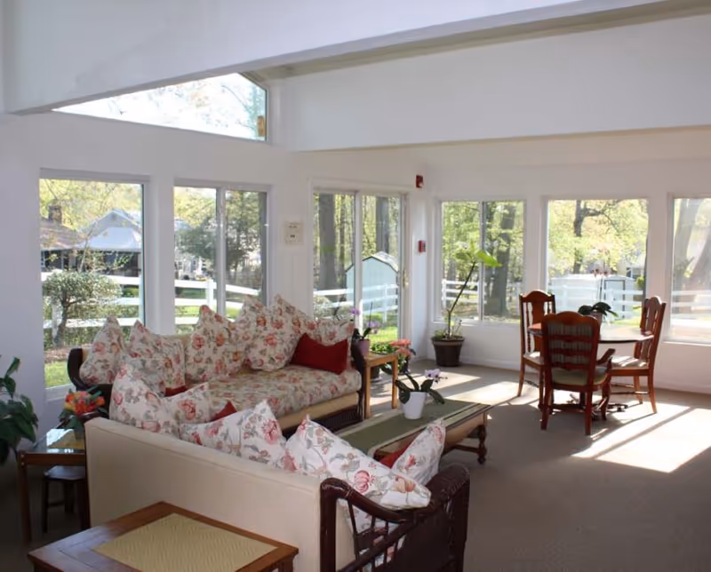 Sunlit common living room with floral sofas, a coffee table, and a dining table surrounded by large windows overlooking trees.