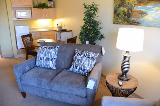 Small living area with a gray loveseat and patterned pillows, side table with lamp, and a kitchenette with dining table in the background.