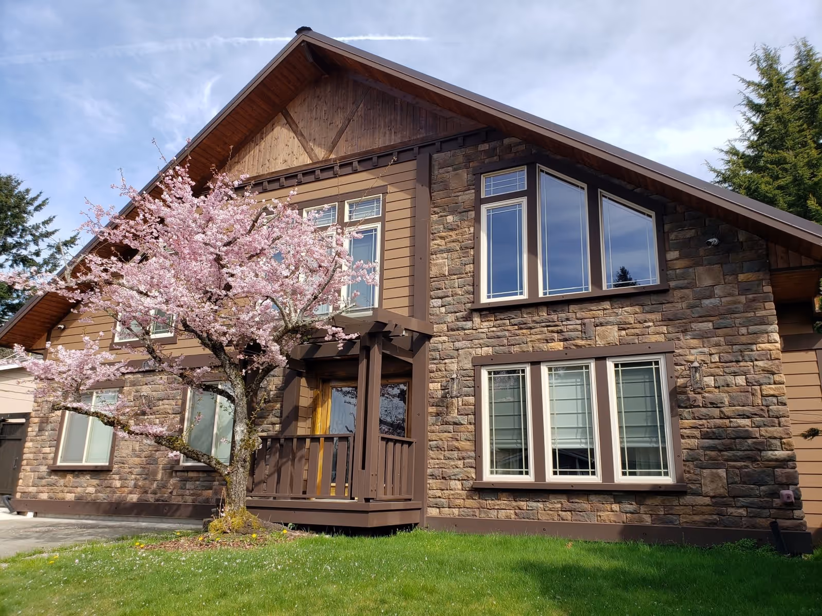 Exterior view of a two-story house with stone and wood siding, featuring multiple windows and a small porch. A blooming pink cherry blossom tree is in the front yard with green grass and a partly cloudy sky in the background.