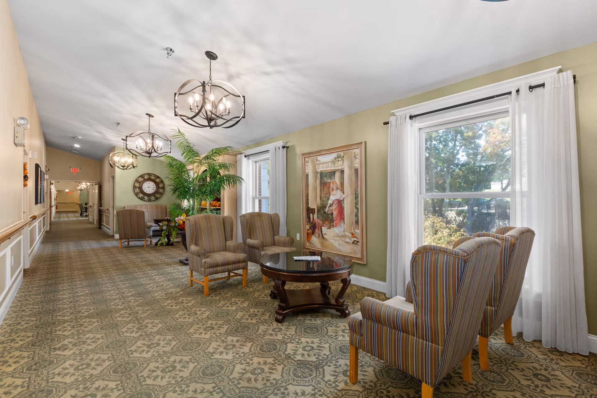 A well-lit sitting area in a hallway of Twin Oaks Estate featuring four striped armchairs arranged around a round wooden coffee table. The walls are painted light green with white trim, and there are two large windows with white curtains letting in natural light. A large framed painting hangs on the wall, and a tall green plant is positioned near the seating area. The hallway extends into the distance with additional seating and decorative elements, including a large wall clock and hanging light fixtures.