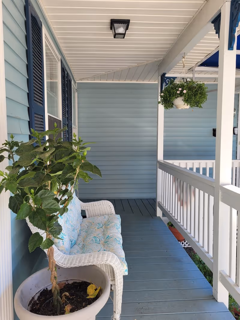 A covered porch area with light blue wooden siding and white railing. There is a white wicker bench with a patterned cushion, a potted plant with green leaves next to the bench, and a hanging basket with green foliage and small pink flowers. The porch floor is painted blue and there is a ceiling light fixture above.