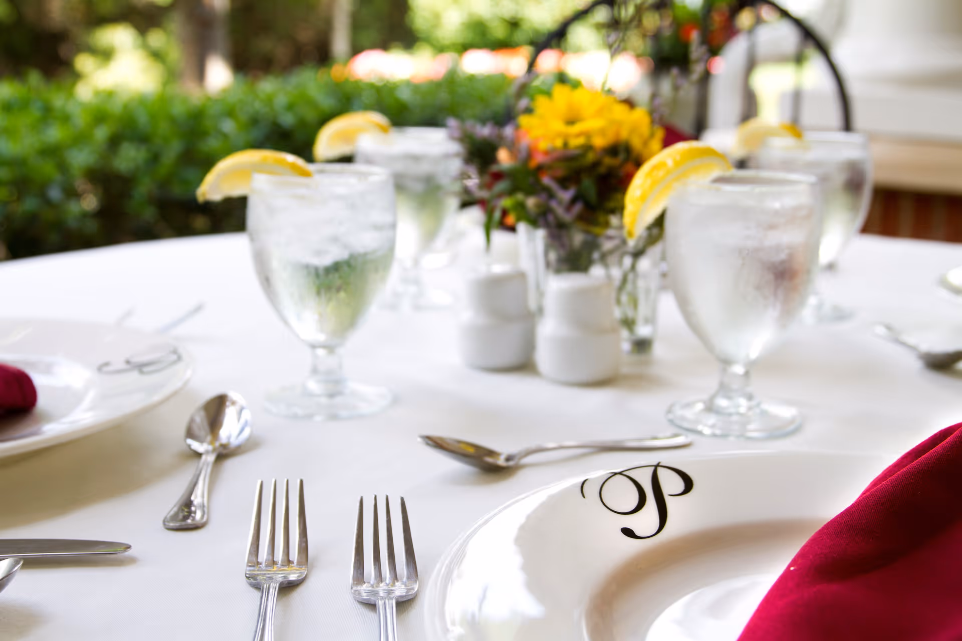A close-up view of a dining table set with white plates featuring a decorative letter 'P', silverware, glasses of water with lemon wedges, and a centerpiece with yellow and orange flowers. The table is covered with a white tablecloth and a red napkin is visible on the right side. The background shows greenery and an outdoor setting.