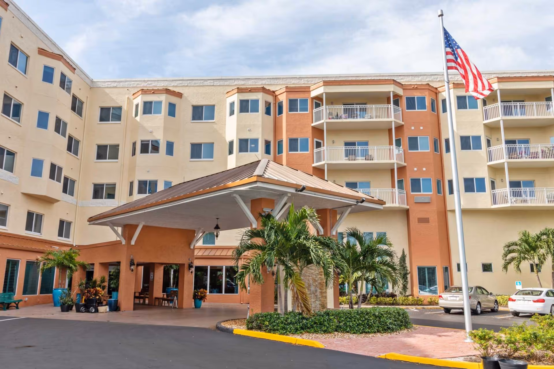 Exterior view of The Palms of Fort Myers senior living facility showing a multi-story building with beige and orange walls, multiple windows and balconies, a covered entrance with a brown roof, palm trees, and an American flag on a flagpole in the parking area.