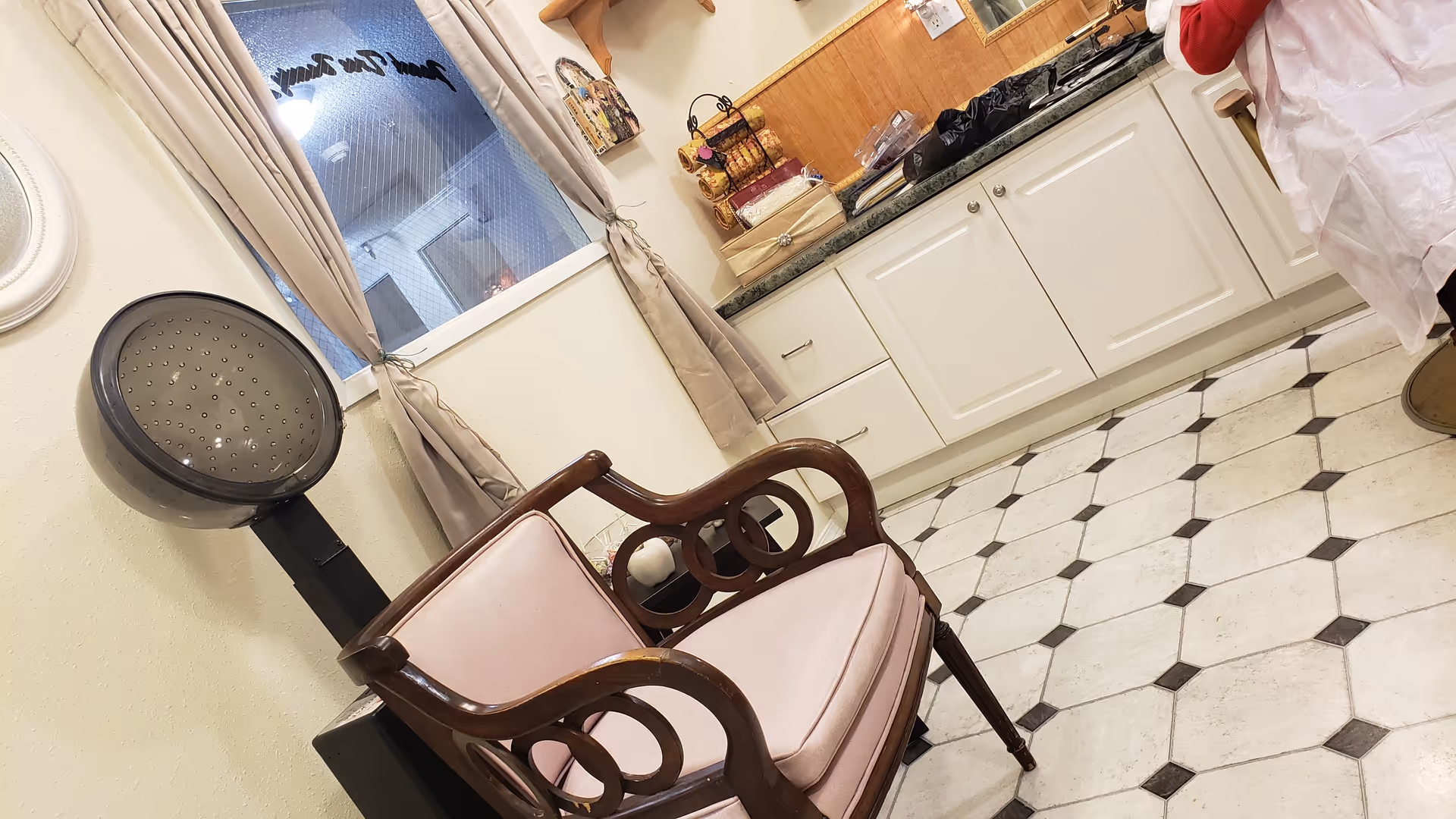 Interior of a salon area in a retirement center featuring a vintage hair dryer, a wooden chair with pink cushions, a window with beige curtains, and white cabinets with a green countertop. The floor has a white and black tile pattern.